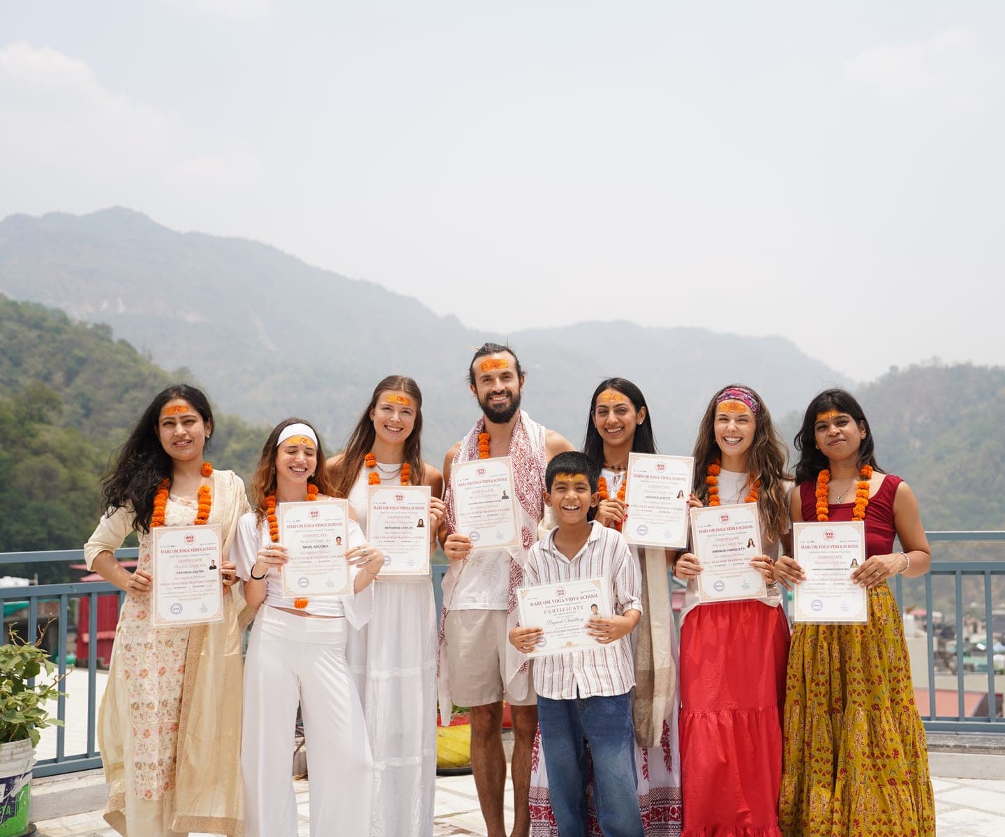 Graduating students of Hariom Yoga Vidya School holding their teacher training certificates, with the Himalayan foothills of Rishikesh behind them